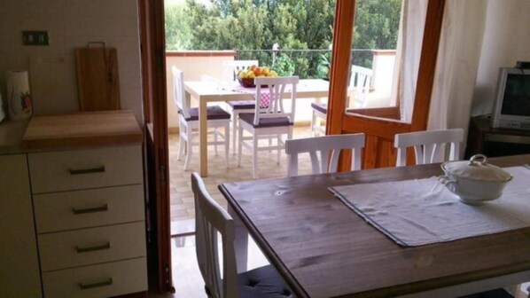 A room with a wooden table, white chairs, a small kitchen area, and a balcony with a view of trees. - Large cozy house 400 meters from the sea ideal for families and groups (Santa Maria Navarrese)