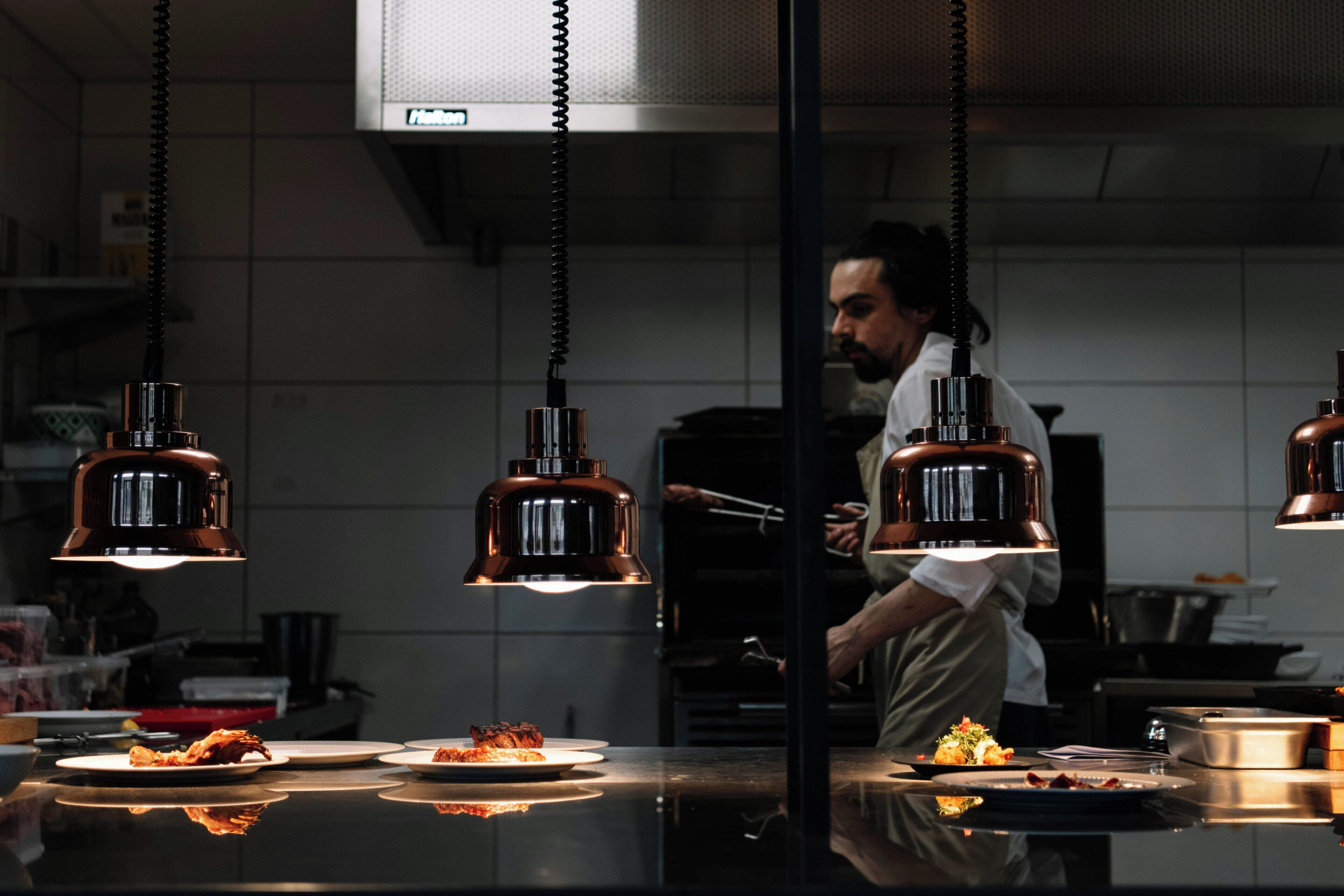 A kitchen with hanging pendant lights, a chef preparing food, and plates of food on the counter.
