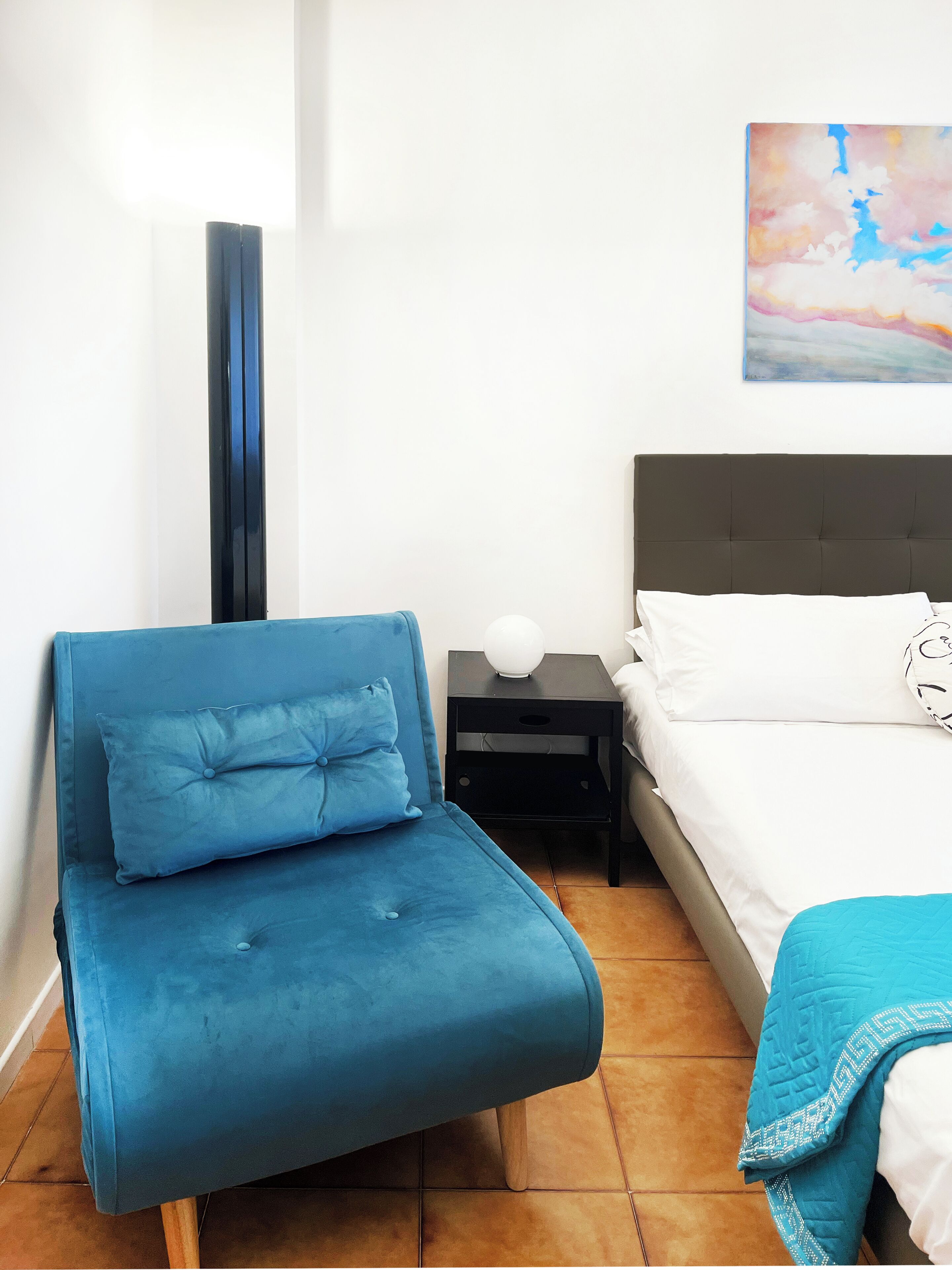 A bedroom with a bed, a blue chair, a bedside table, and a wall painting.