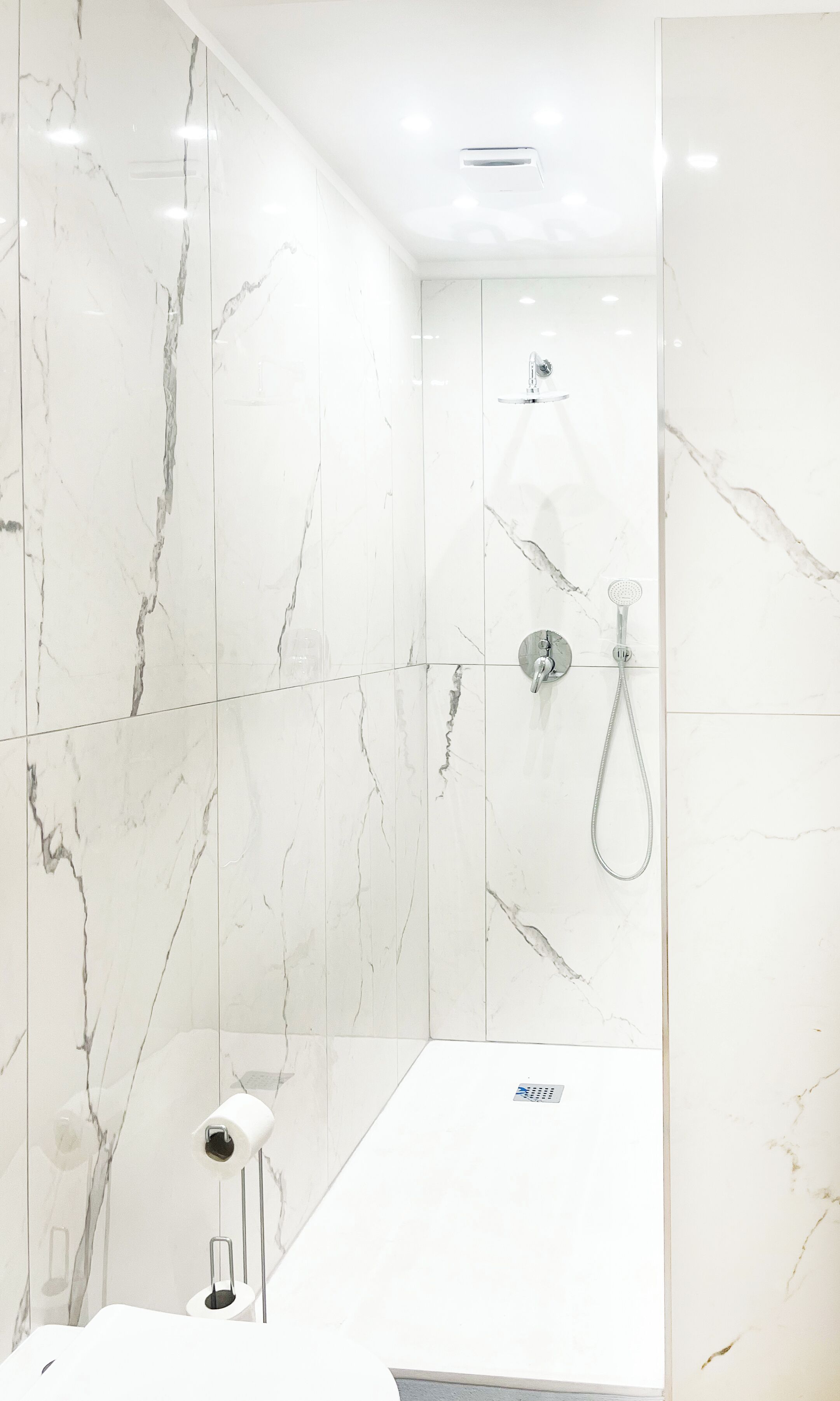 A modern bathroom with white marble walls, a shower with a handheld showerhead, and a white sink with a mirror.
