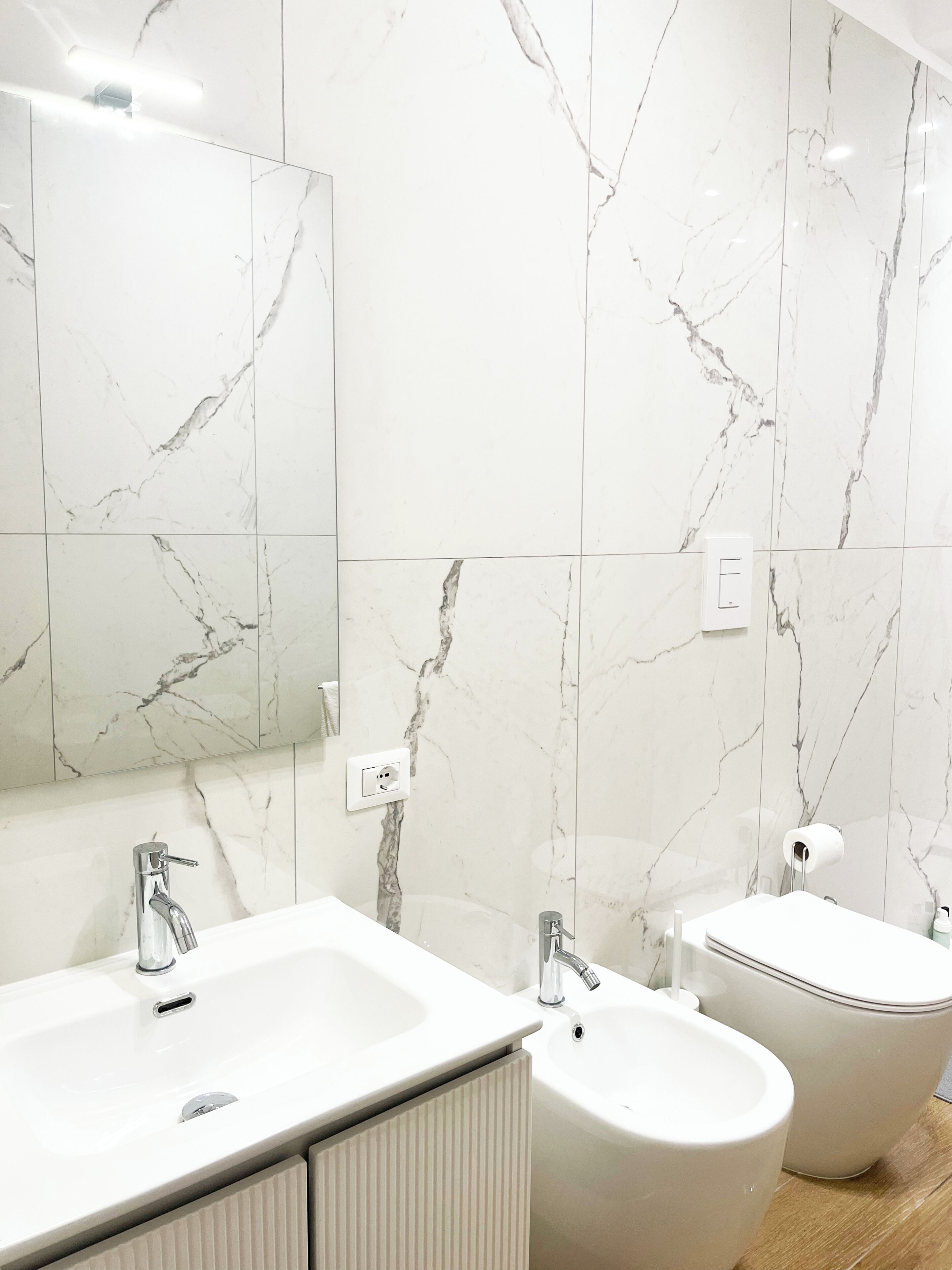 A modern bathroom with marble walls, a large mirror, two sinks, and a bidet.