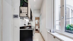 Une cuisine étroite avec un réfrigérateur noir, des placards blancs et une fenêtre donnant sur l’extérieur.