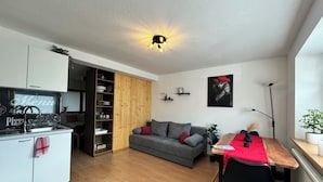 A compact living space with a kitchenette, a grey sofa, a dining table with a red tablecloth, and a wooden floor.