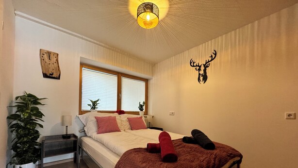 A bedroom with a bed, a nightstand, a potted plant, a wall clock, and a window with blinds.