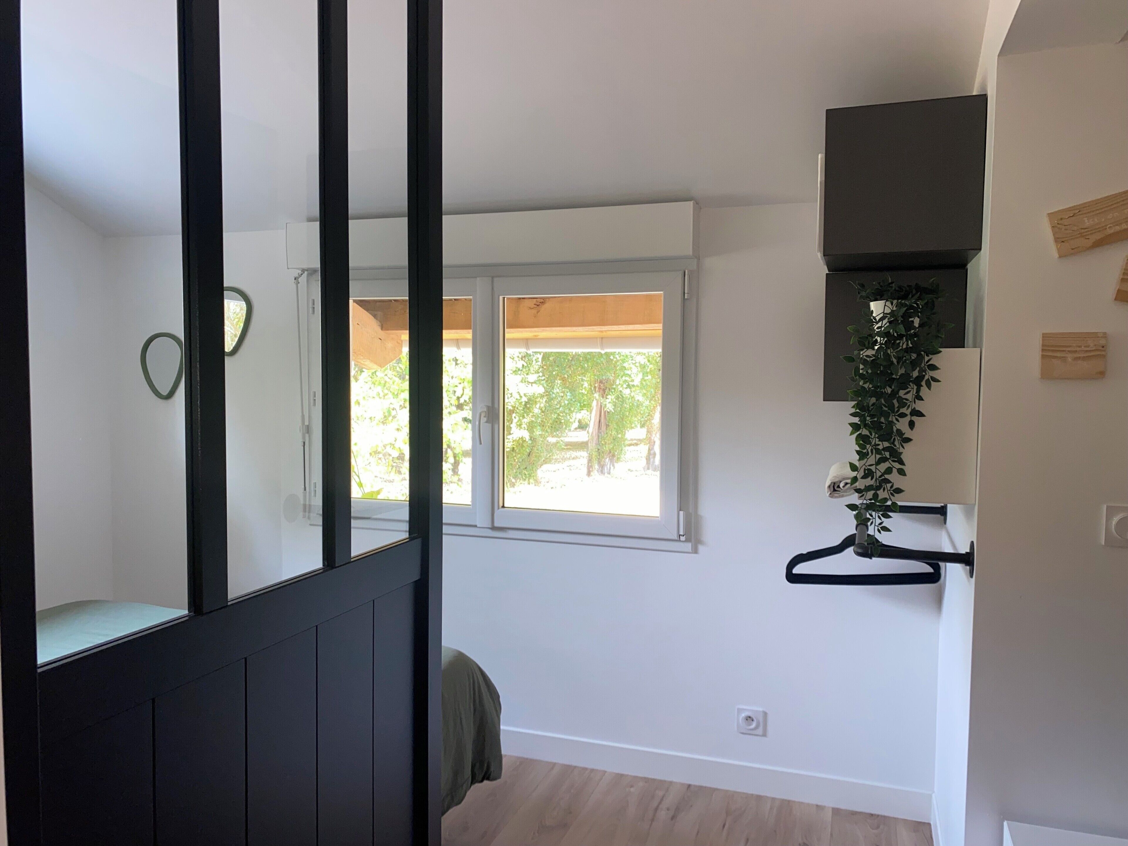 Une chambre avec un lit, une fenêtre donnant sur un espace verdoyant, un téléviseur fixé au mur et une plante fixée sur un autre mur.