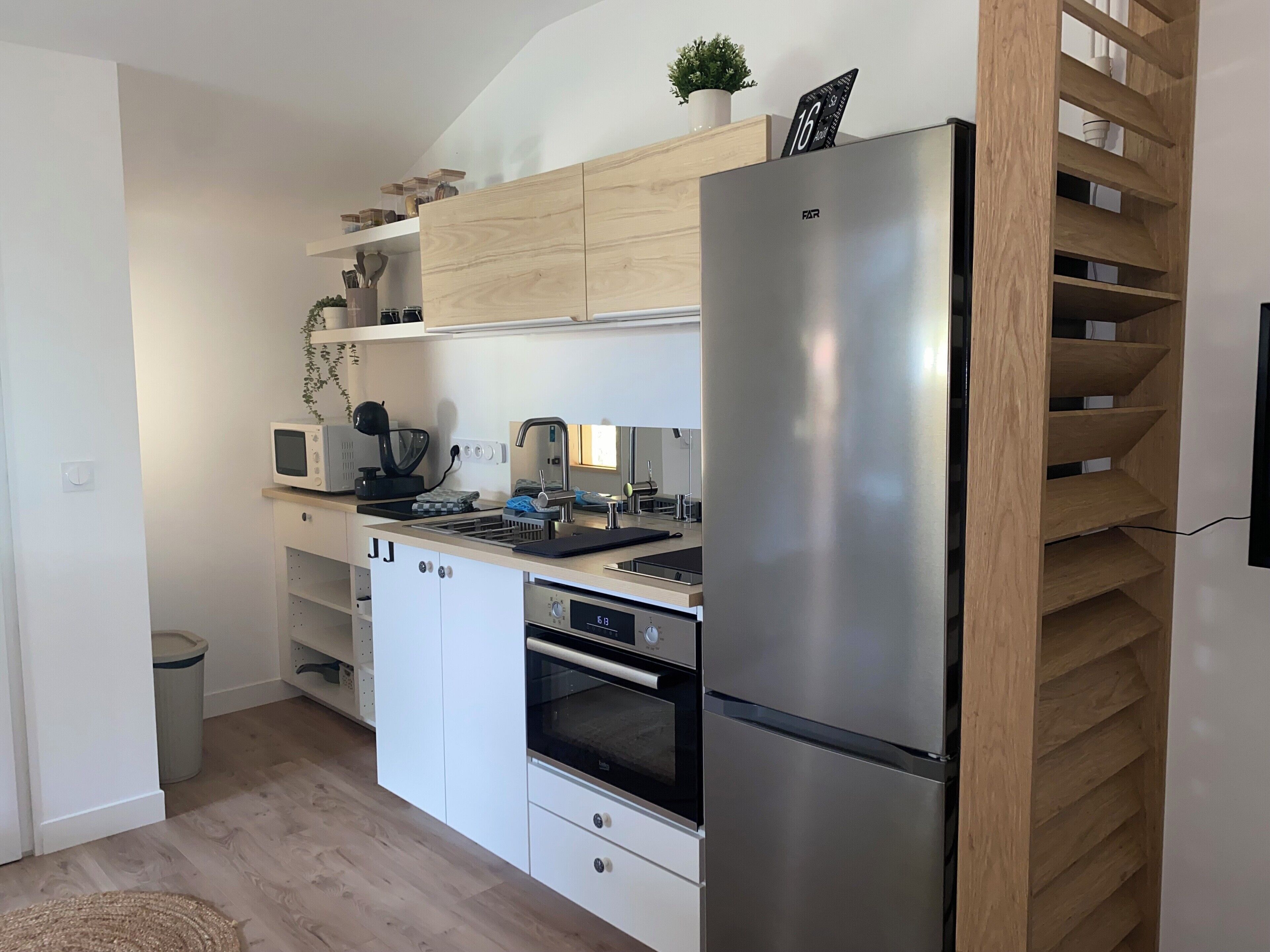 A compact kitchen with a stainless steel refrigerator, a microwave, a kettle, and a built-in oven. There are wooden shelves above the counter and a small potted plant on the wall.