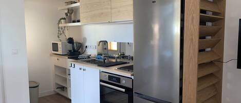 A compact kitchen with a stainless steel refrigerator, a microwave, a kettle, and a built-in oven. There are wooden shelves above the counter and a small potted plant on the wall.