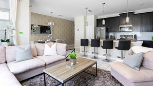 A modern living room with a sofa, coffee table, and two chairs. A kitchen with dark cabinets and a microwave is visible in the background. - Storey Lake 4BR Home - Private Pool (Kissimmee)