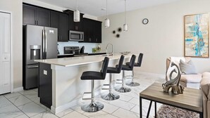 A modern kitchen with dark cabinets, stainless steel appliances, a white island with bar stools, pendant lighting, and a decorative piece on the coffee table. - Storey Lake 4BR Home - Private Pool (Kissimmee)