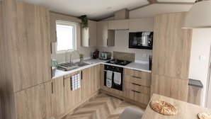 A modern kitchen with wooden cabinets, a microwave, a stove, and a sink. - The Beach Hut, Burghead Holiday Park (Burghead)