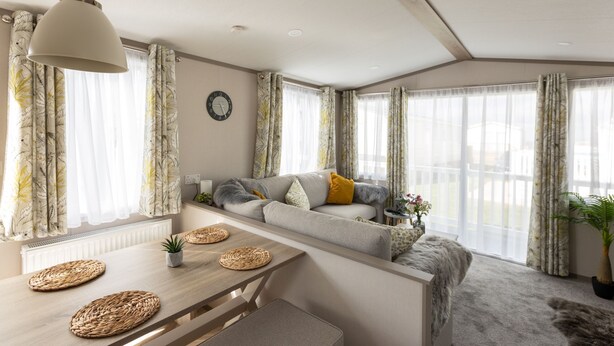 A modern living room with a sofa, a dining table with placemats, and large windows with sheer curtains. - The Beach Hut, Burghead Holiday Park (Burghead)