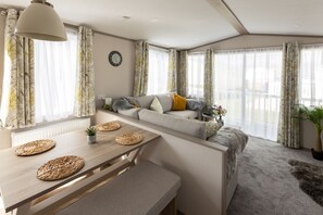 A modern living room with a sofa, a dining table with placemats, and large windows with sheer curtains. - The Beach Hut, Burghead Holiday Park (Burghead)