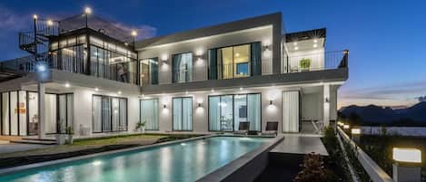 A modern multi-story house with a large swimming pool, multiple balconies, and outdoor lighting.