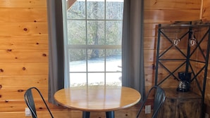 A wooden cabin interior with a round wooden table and two black chairs. A window with a view of trees and a gravel path is visible. - Red Cozy Cottage  1-bedroom cottage in charming Oneida, TN (Oneida)