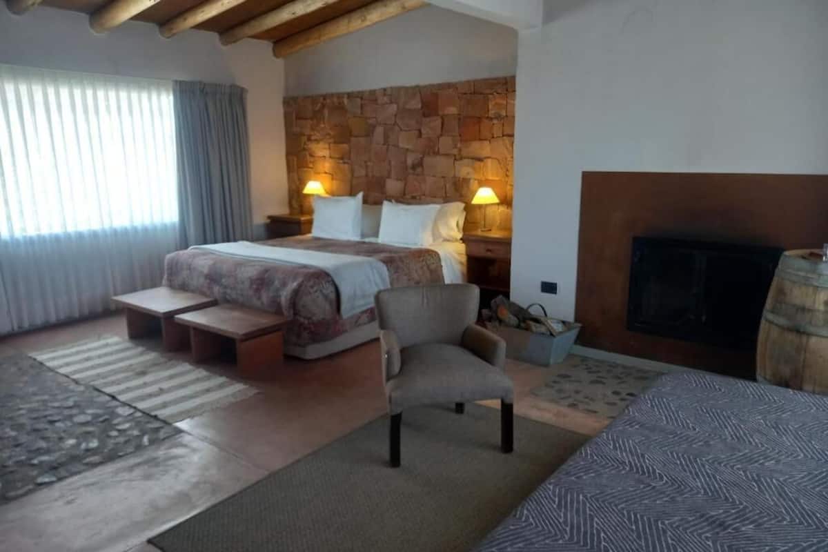 A bedroom with a stone headboard, a bed with two bedside tables, a chair, a fireplace, and a window with curtains.