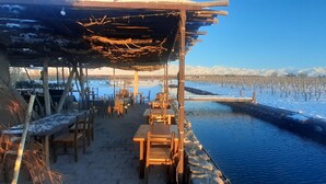 A riverside outdoor seating area with tables and chairs, a wooden pergola overhead, and a snow-covered landscape with mountains in the background. - Tupungato Divino (Tupungato)