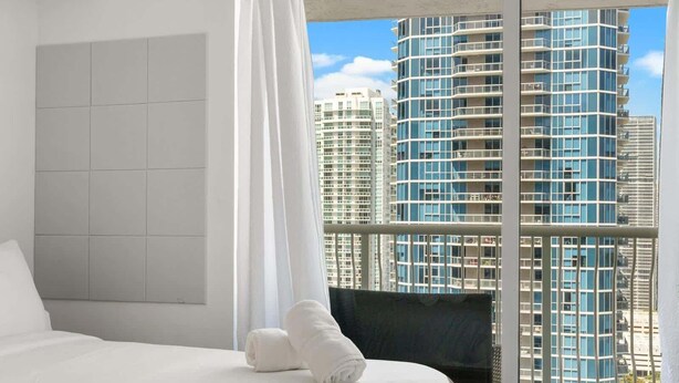 Habitación de hotel con una cama grande, vista a rascacielos y un balcón con barandilla de vidrio.