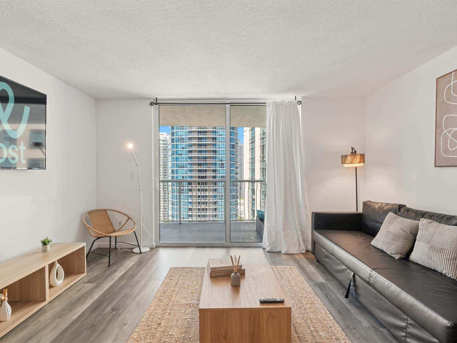 A modern living room with a sofa, a chair, a coffee table, and a view of tall buildings through a glass door.