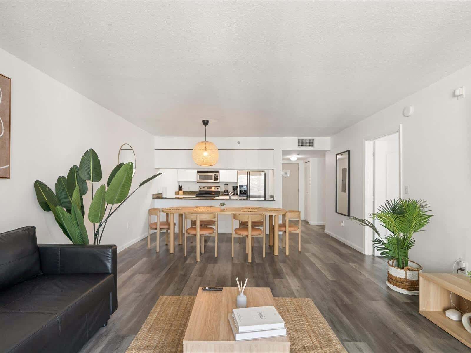 A modern living room with a black sofa, a dining area with wooden chairs, a kitchen with white cabinets, and a large plant.