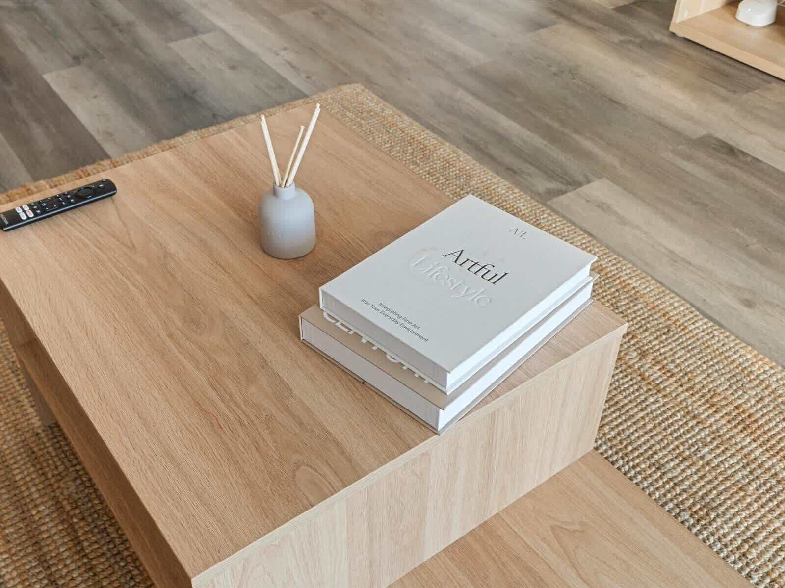 A wooden coffee table with a remote control, a white book titled 'Artful Lifestyle,' and a small vase with reed sticks.