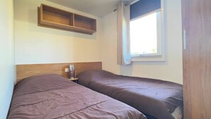 A bedroom with two beds, a wooden headboard, a bedside table with a lamp, and a window with curtains. - 6 person mobile home "La Pagode" in a family campsite**** (Fréjus)