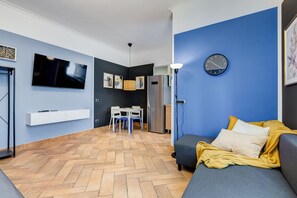 Ein modernes Wohnzimmer mit Sofa, Fernseher, Esstisch mit Stühlen, Küchenzeile und einer Uhr an der Wand.