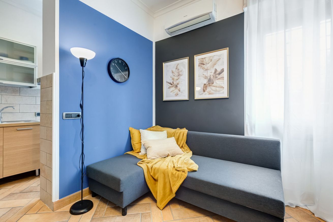 A modern living room with a grey sofa, a blue accent wall, a floor lamp, a clock, and two framed botanical prints.