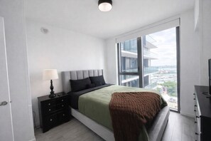 Ett modernt sovrum med en säng, ett sängbord, en lampa och ett stort fönster med utsikt över staden.