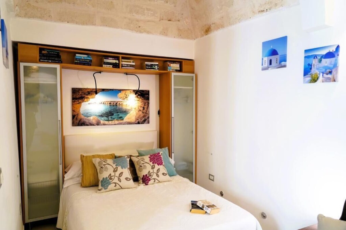 A bedroom with a bed, a nightstand, a wall-mounted TV, and a shelf with books.