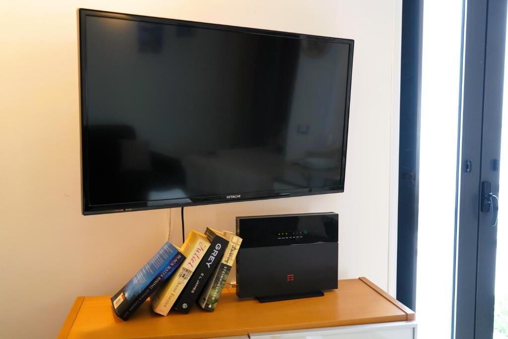 Ein flachbildfernseher an der Wand, ein Holztisch mit Büchern und einem schwarzen elektronischen Gerät, und eine Tür zur rechten Seite.