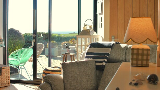 A cozy living room with a large sliding glass door leading to an outdoor deck, a comfortable sofa, a lamp, and a small table with decorative items. - Mill View Cottage- charming coastal cottage (Saint Monans)