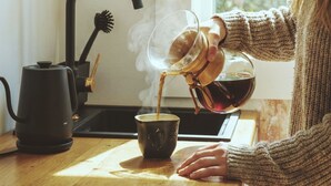 Iemand giet thee in een kop aan een keukentafel, met een waterkoker en een koffiezetapparaat.