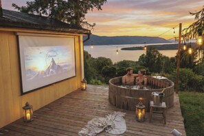 Une terrasse en bois avec un jacuzzi, un écran de projection diffusant un paysage de montagne et une vue sur le lac.