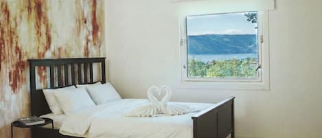 Cama com colcha e almofadas brancas, cabeceira de madeira, mesa de cabeceira pequena com um livro e vista para montanhas pela janela.
