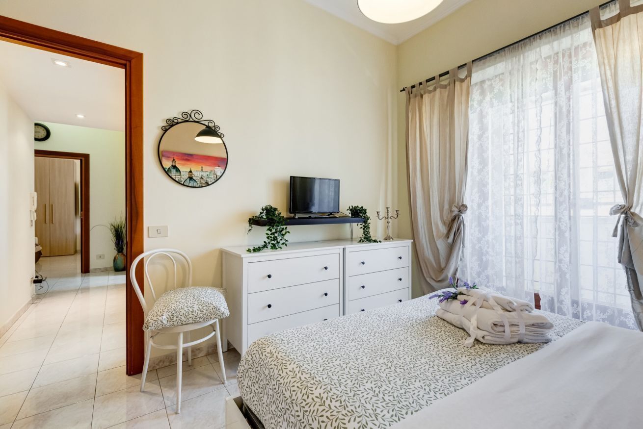 Un dormitorio con cama, televisión, cómoda, silla y ventana con cortinas.