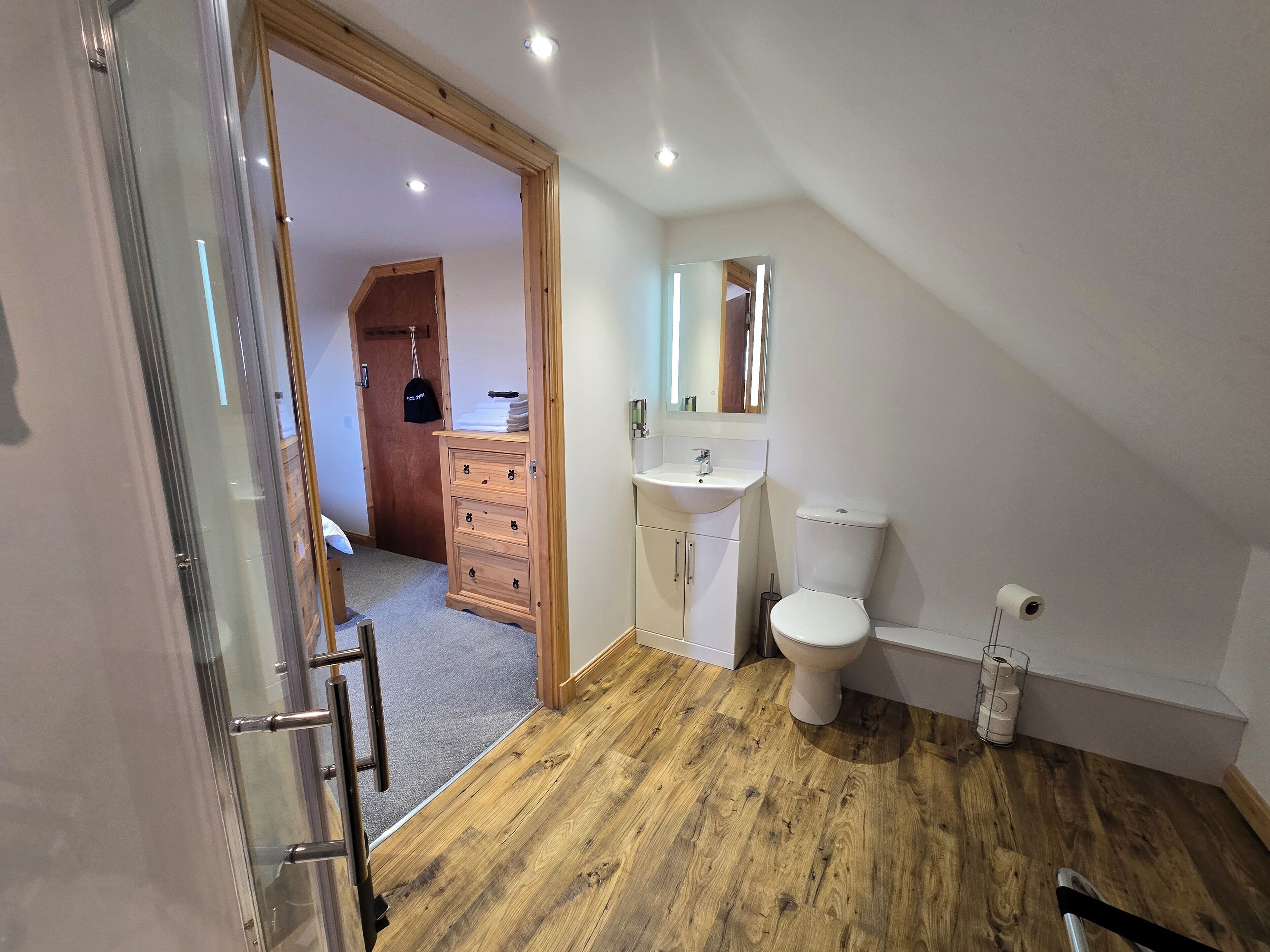 A modern bathroom with a wooden floor, a white sink, a toilet, and a glass shower enclosure.