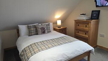 Ein ordentlich bezogenes Bett mit einer Karo-Decke und Kissen, eine Nachttischlampe, eine Holzkommode mit Schubladen und ein an der Wand befestigtes Fernsehgerät.
