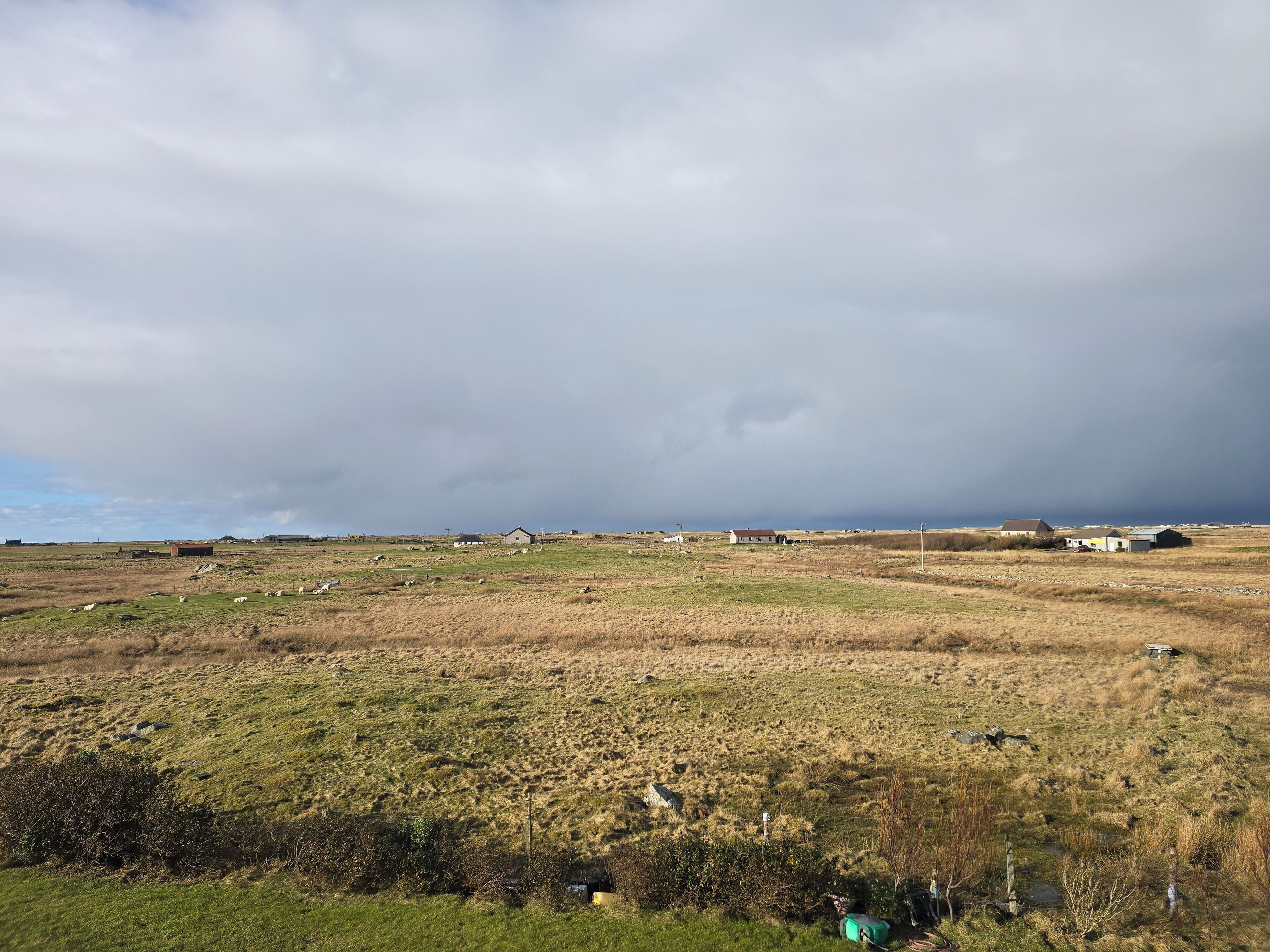 A rural landscape with scattered buildings, a grassy field, and a cloudy sky.
