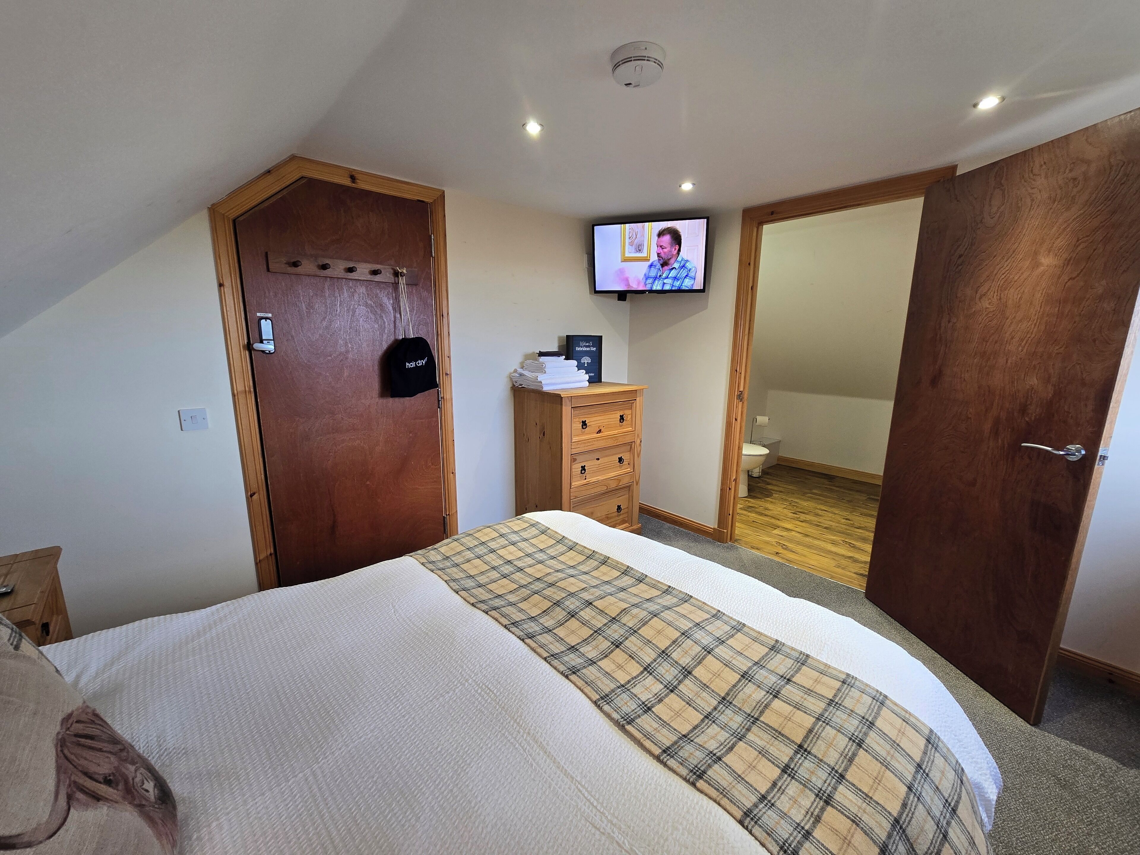 Un dormitorio con una cama, un aparador de madera, un televisor montado en la pared y una puerta que da a otra habitación.