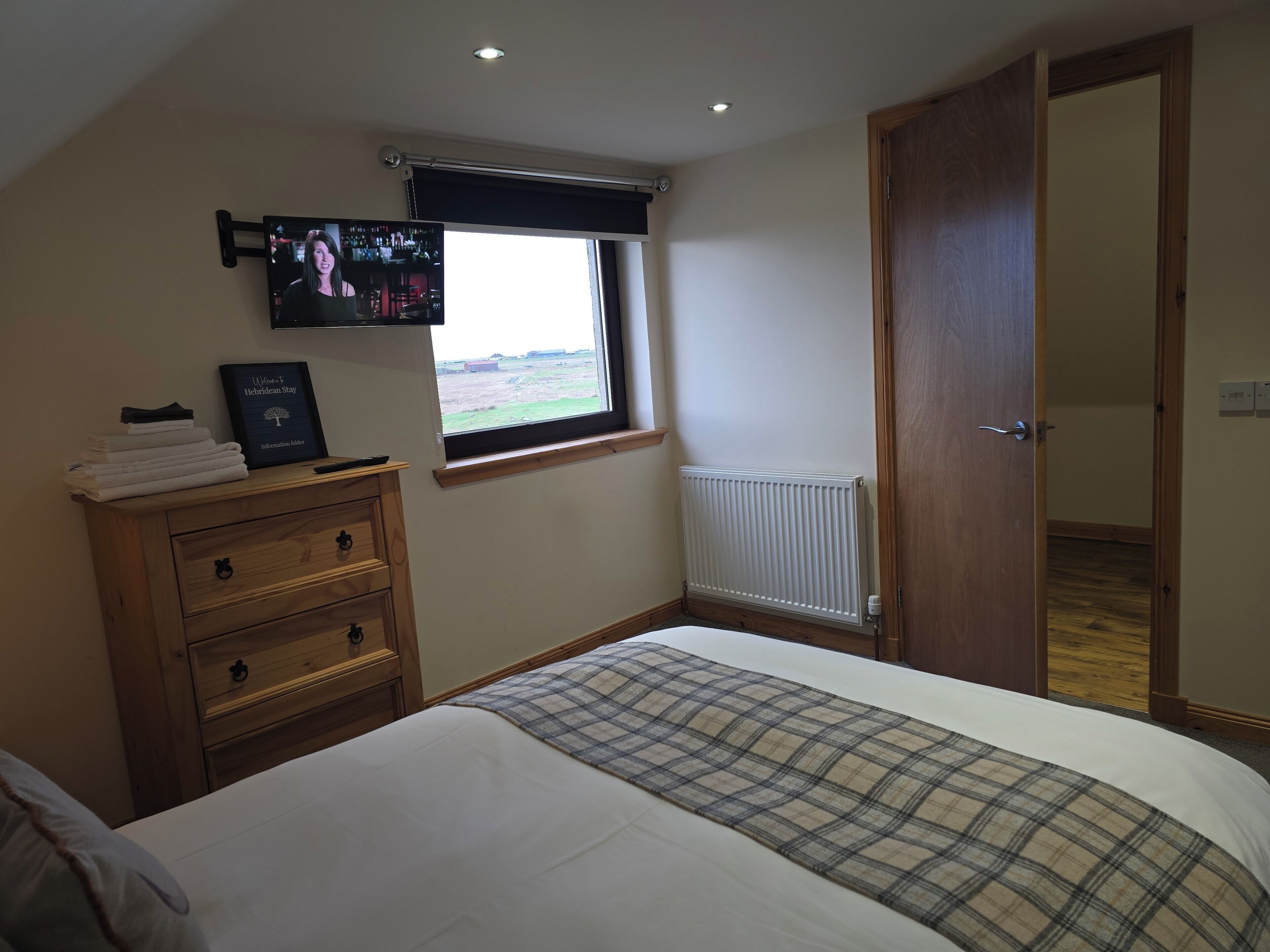A bedroom with a bed, a wooden dresser, a television mounted on the wall, a window with a view of the countryside, and a radiator.
