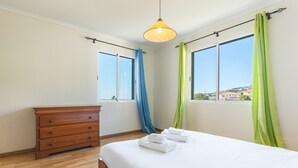 A bedroom with a wooden dresser, a bed with white linens, and a view of the city through large windows. - Sunrising Apartment by Atlantic Holiday (Santa Cruz)