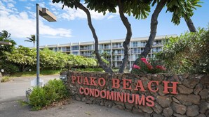 Un mur de pierre sur lequel on peut lire l’inscription « PUAKO BEACH CONDOMINIUMS », entouré de verdure et de palmiers.