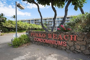 Un muro de piedra con el letrero "PUAKO BEACH CONDOMINIUMS", rodeado de follaje y palmeras.