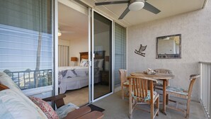 Un balcon doté d’un ventilateur de plafond, une table à manger avec des chaises, et un lit donnant sur une vue à travers une porte-fenêtre coulissante.