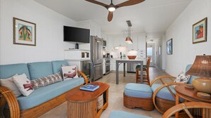 Un salon moderne avec un canapé, une table basse et des fauteuils en osier. On aperçoit, au fond, une cuisine avec une table à manger et des chaises.