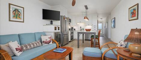 Una sala de estar moderna con un sofá, una mesa de centro y sillas de mimbre. Se ve una cocina con una mesa de comedor y sillas al fondo.