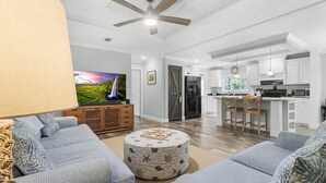 A modern living room with a large flat-screen TV, a ceiling fan, and a kitchen area with white cabinets and a bar stool. - Fin & Feather - Steinhatchee Florida (Steinhatchee)
