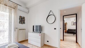 Une chambre d’hôtel avec un lit, un téléviseur posé sur une commode, un radiateur, un grand miroir rond fixé au mur et une porte donnant sur une autre pièce.