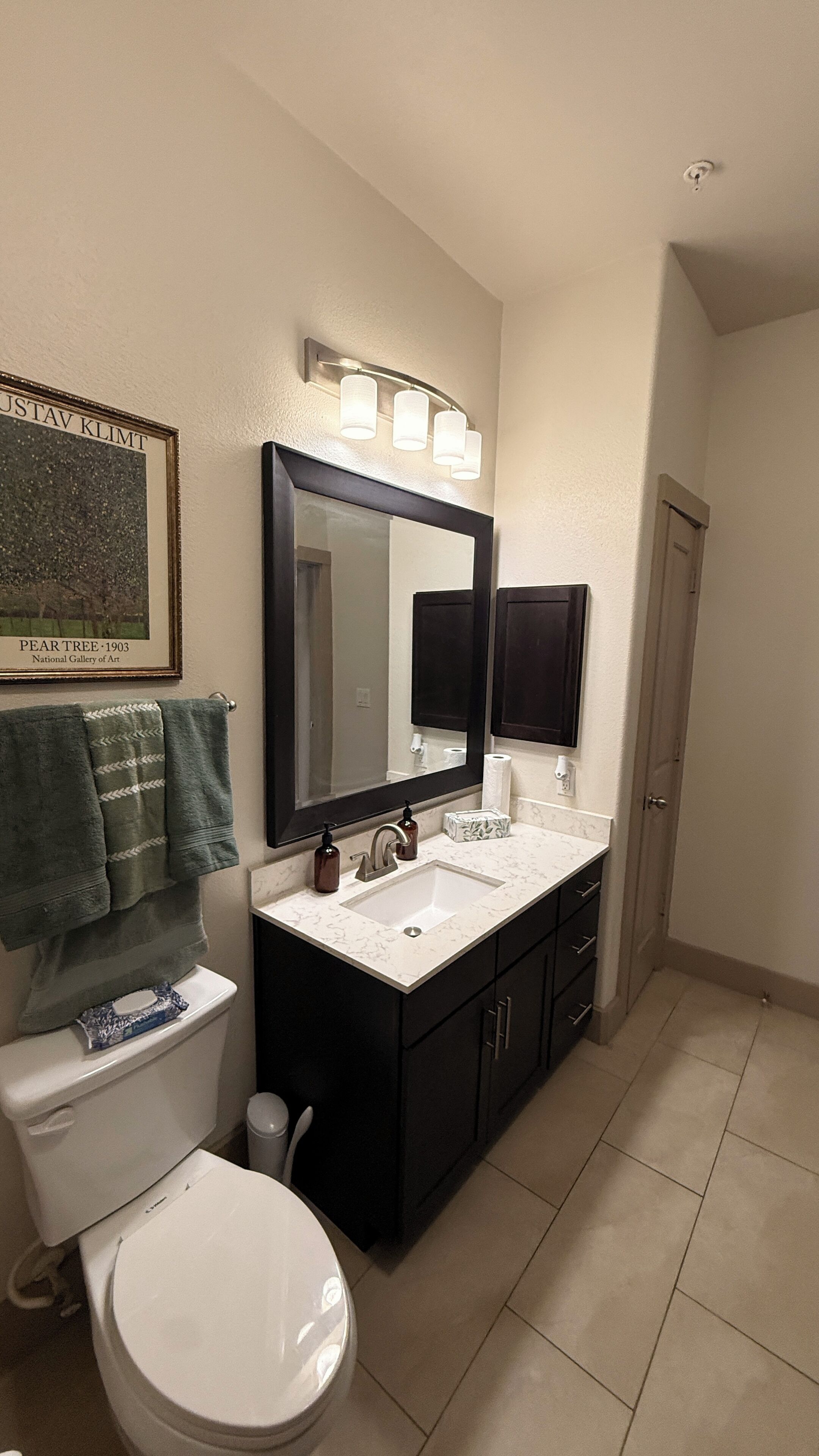 A bathroom with a toilet, sink, and mirror. There is a framed artwork above the sink and a towel hanging on the wall.