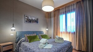 A modern bedroom with a large bed, two bedside lamps, a cityscape painting, and a view of the outdoors through sheer curtains. - Accogliente appartamento situato a Olbia,
A 2 km dall'aeroporto.. (Olbia)